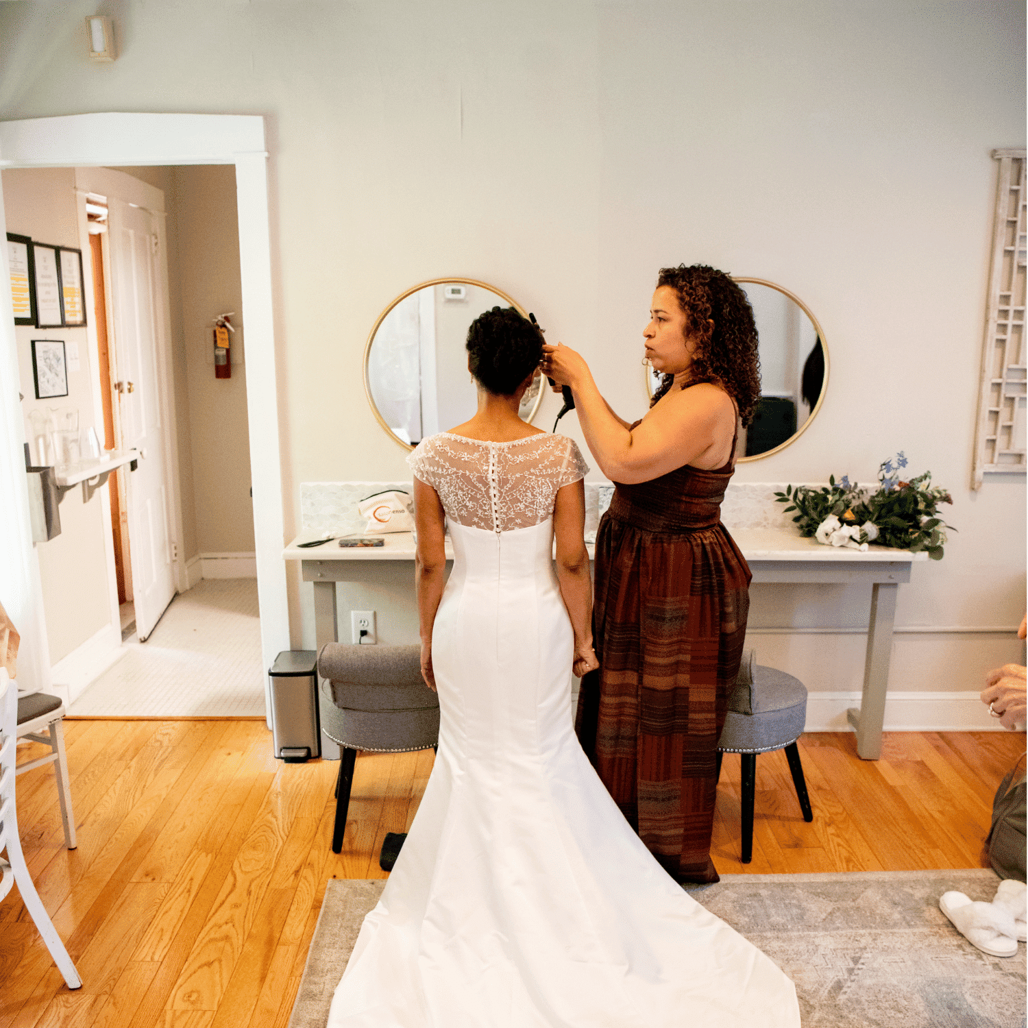 Bride getting ready in elegant white dress with hair styled by a woman in a warmly lit room.